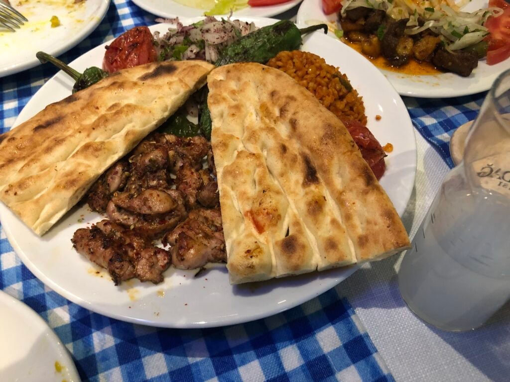 Plate of mixed grill with grilled meat pieces, two flatbreads, peppers, and yellow rice on a blue-checkered tablecloth, in a dining setting.