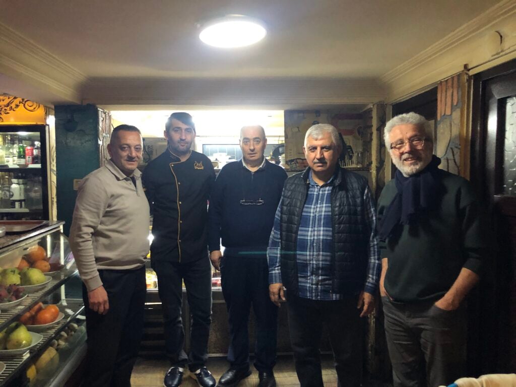 Five men stand together inside a small shop or cafe, with a fruit display on the left and warm lighting behind them.