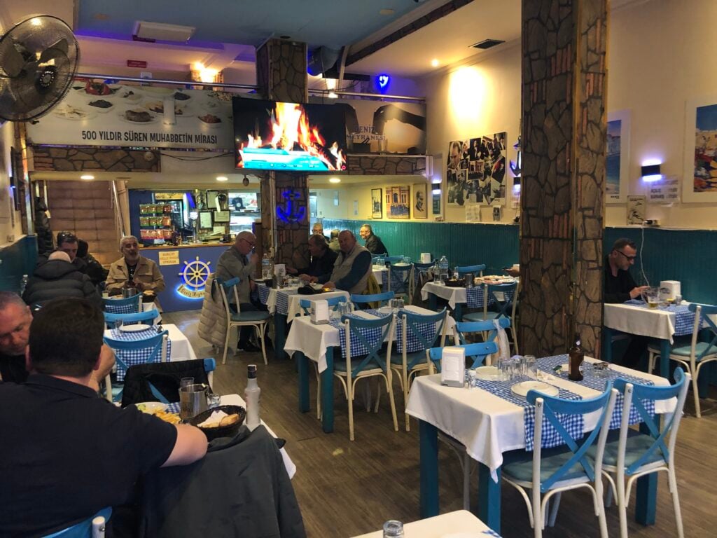 Restaurant dining hall with blue chairs and checkered tablecloths, diners seated at tables, a wall-mounted TV showing a flame, and stone pillars in the background.