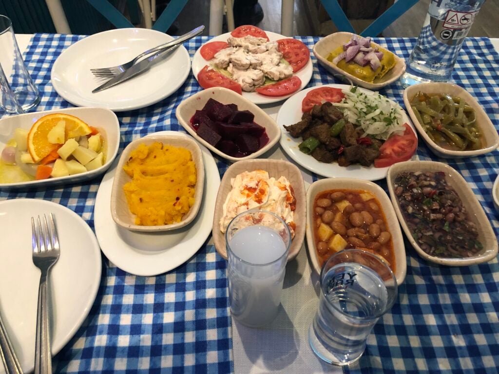 Table of assorted dishes on a blue-check tablecloth, including beet salad, chickpeas, orange wedges, and tomato slices around a chicken/tuna salad plate.