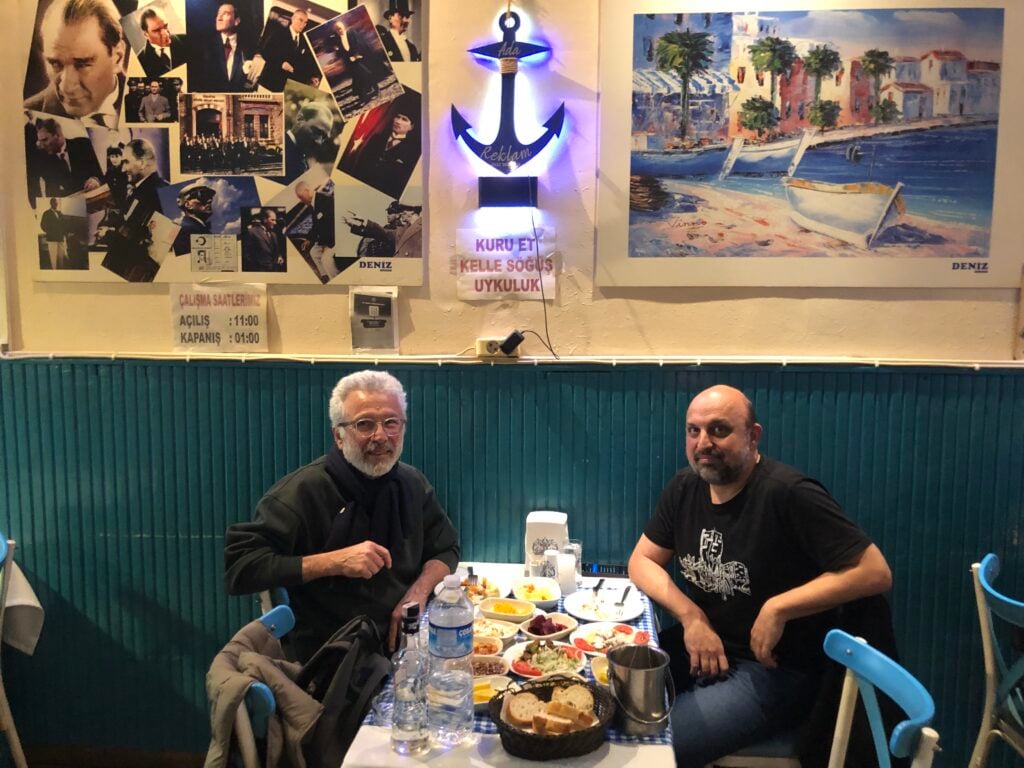 Two men sit at a restaurant table with plates of food, water, and bread, smiling at the camera; wall posters and a large blue anchor sign decorate the background.