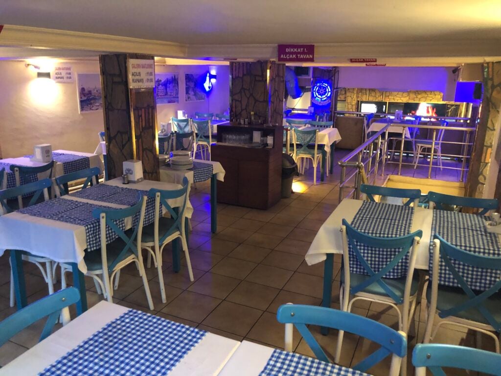 Diner-style restaurant interior with blue chairs, white tables, and blue-checkered tablecloths; bar area with blue lighting in the back.