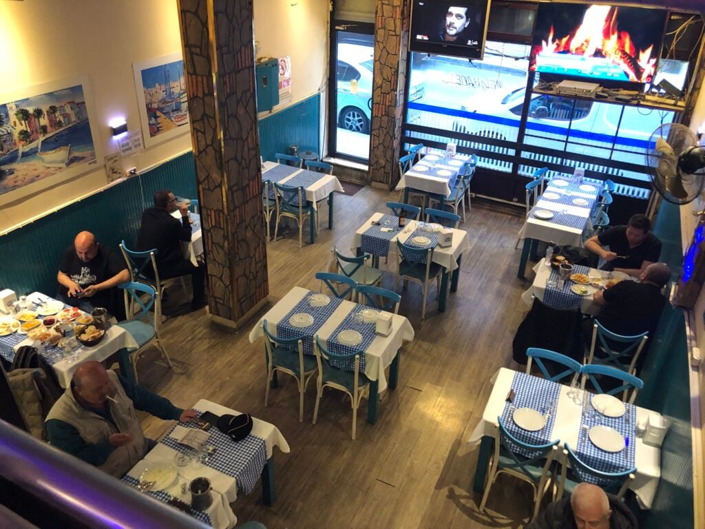 Restaurant dining area with blue chairs and blue-and-white checkered tablecloths; diners seated at scattered tables.