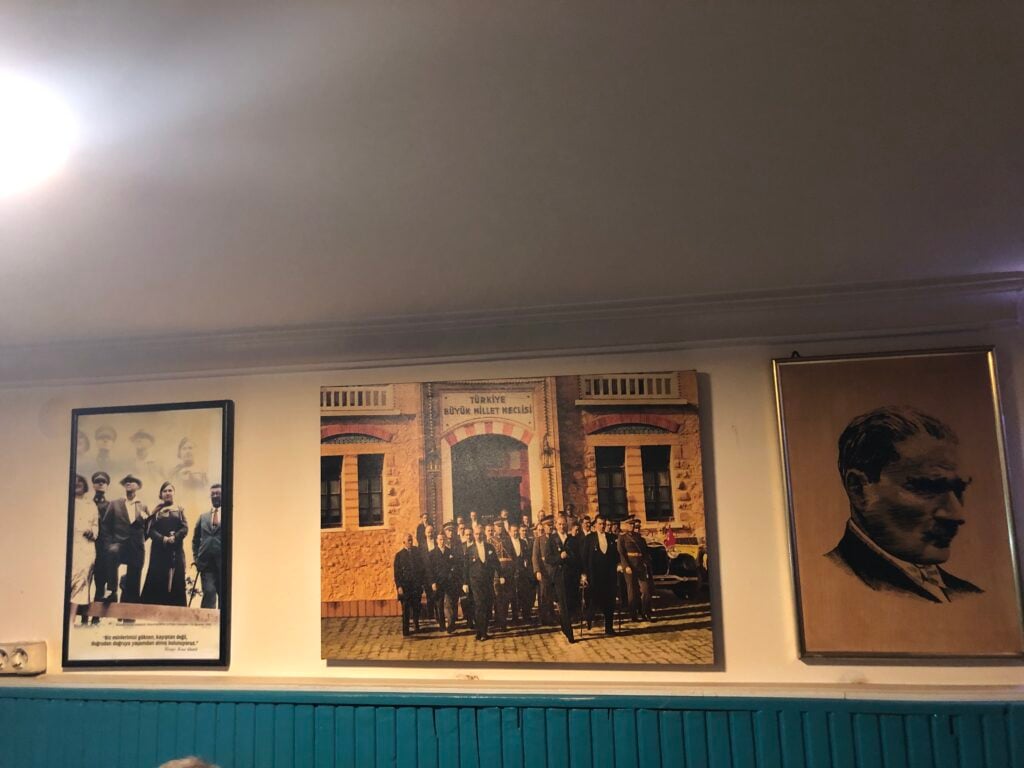 Three framed images hung on a wall: left a black‑and‑white group photo, center a large group photo outside a brick building, right a sepia portrait of a man.