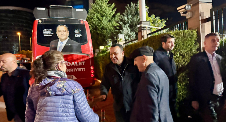 Nighttime street scene with a red campaign bus displaying a politician's portrait and a group of people greeting each other nearby.