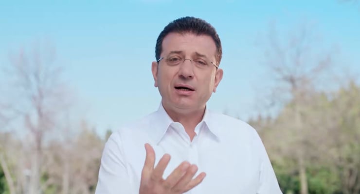 Man in a white shirt speaks to the camera outdoors, with a blue sky and trees blurred in the background.