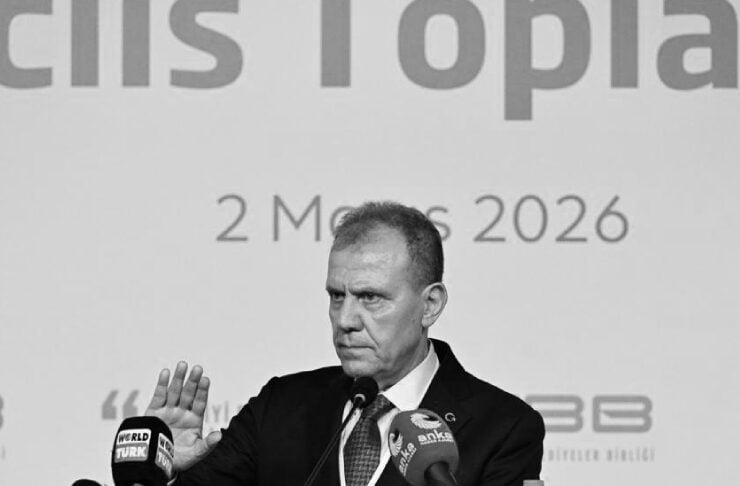 Man in a suit speaking at a podium with multiple microphones, raising his right hand, backdrop shows '2 Mayıs 2026' and logos.
