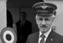 Older man in a pinstripe suit and captain's hat holds a circular signal disc near an aircraft door.