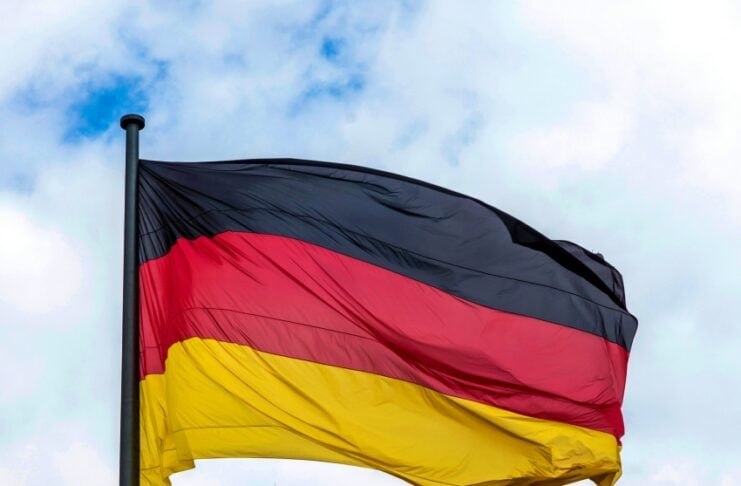 German flag (black-red-gold) fluttering in the wind atop a flagpole against a cloudy sky, with a historic stone monument beneath.