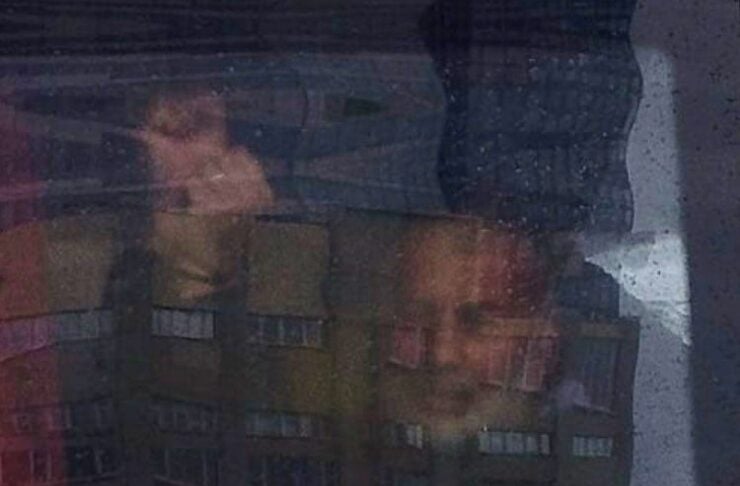 Reflection of a multi-story apartment building on a glass surface, with a faint blurred face superimposed over it.