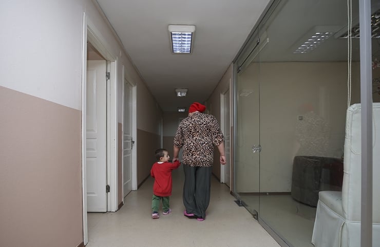 Child in a red shirt holding hands with an adult in a leopard-print top walking down a long hallway with white doors and fluorescent lights on the ceiling.