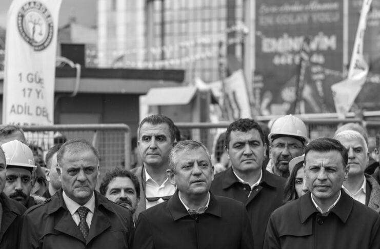 Group of suited officials and workers at a press conference behind a row of microphones at a construction site (black and white).