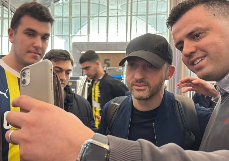 Group of men posing for a selfie with a smartphone in a bright indoor space, one wearing a black cap and backpack on the center-right.