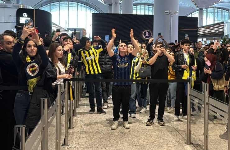 Crowd of fans behind barriers in a bright terminal, many wearing yellow-and-black jerseys and taking photos with their phones.