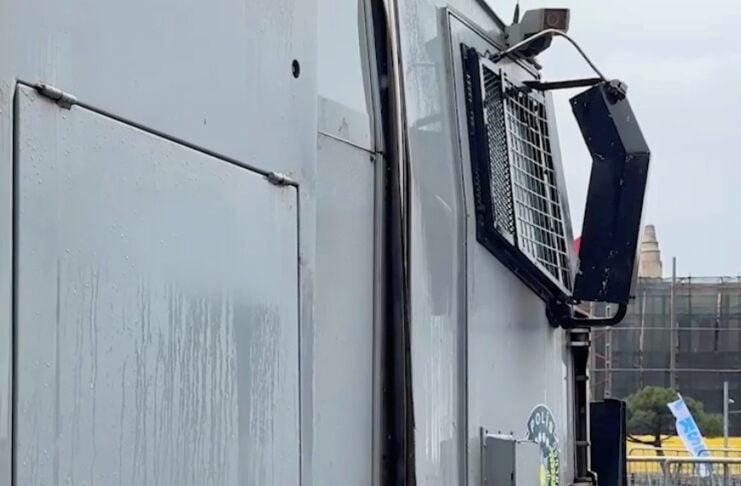Side view of a gray armored police vehicle with a reinforced window grille and a police emblem on the door, parked near a fence.