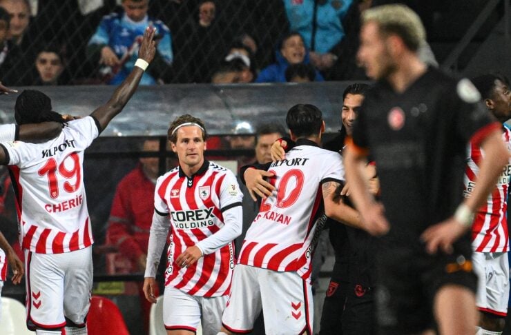 Soccer players in white and red striped uniforms celebrate on the field together after a goal, with fans in the stands behind them.