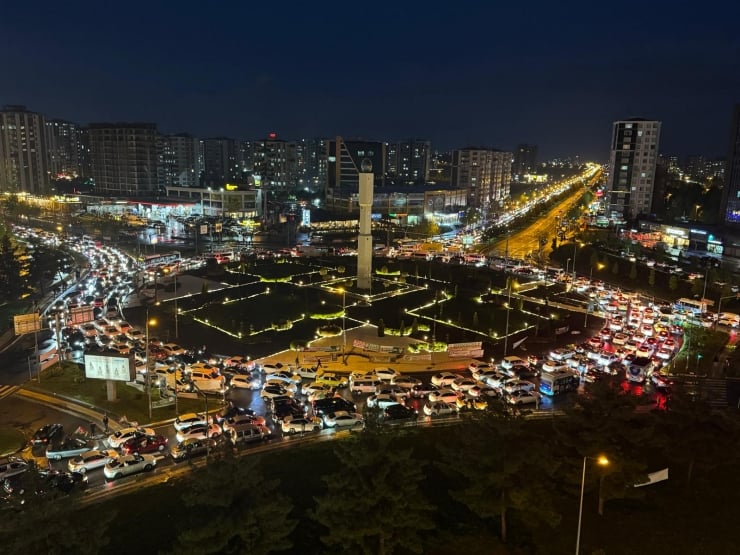 Aerial night view of a large circular roundabout with a tall obelisk in the center, surrounded by heavy city traffic.