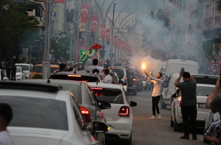 Crowded city street with cars lined up, people waving Turkish flags and a man holding a flare amid smoke.