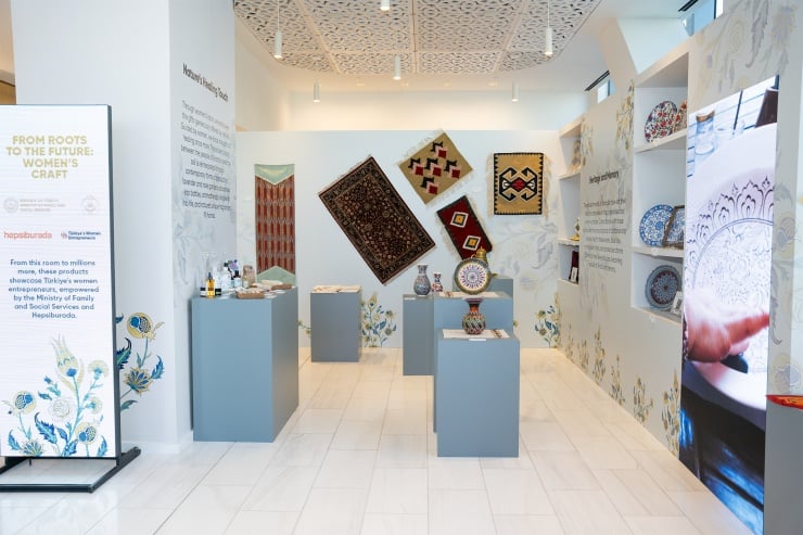 Gallery showroom with woven carpets hung on the back wall and ceramic vases on pedestals; white walls, floral accents, and informative panels along the sides.