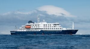 Blue-and-white cruise ship sailing on calm ocean with orange lifeboats on deck.