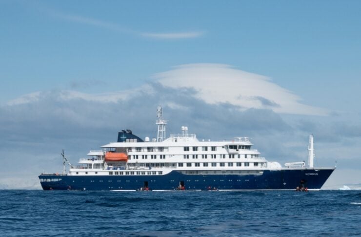 Large blue-and-white passenger yacht on calm ocean water, horizon line with cloudy sky behind it.