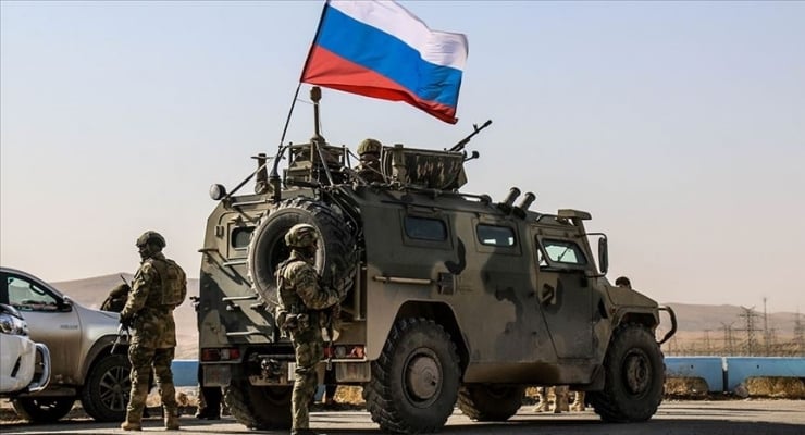 Armored military vehicle with a Russian flag on top, soldiers in camouflage standing nearby on a road in a desert-like area