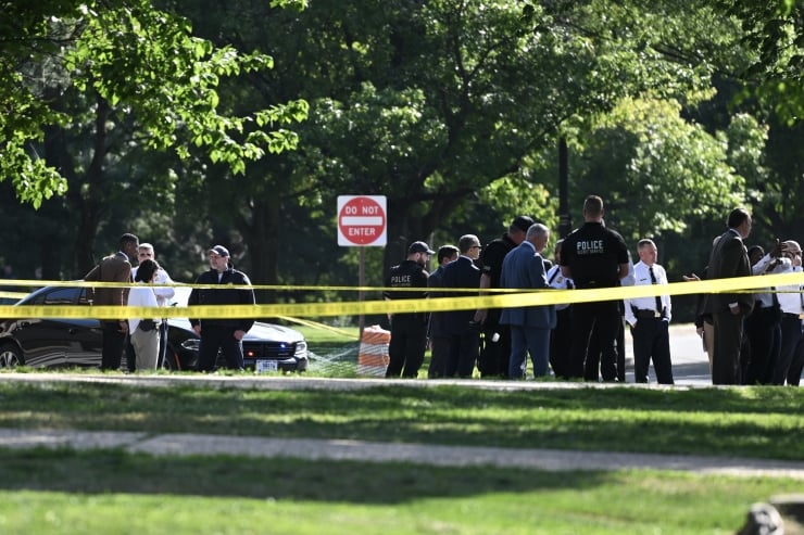 Police officers and investigators standing behind yellow caution tape in a park, with a Do Not Enter sign and cars in the background.