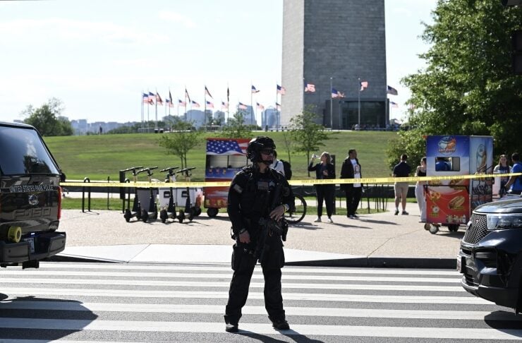 Armed security officer in tactical gear stands at a crosswalk behind yellow caution tape with a monument and flags in the background.