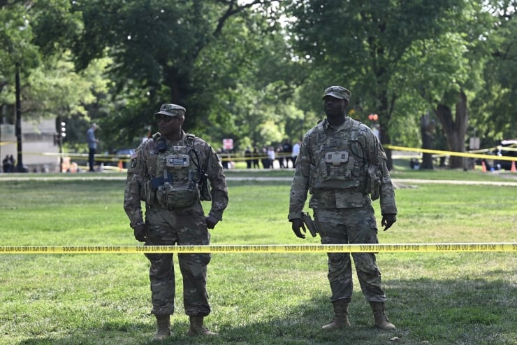 Two soldiers in camouflage uniforms stand in a park behind yellow caution tape.
