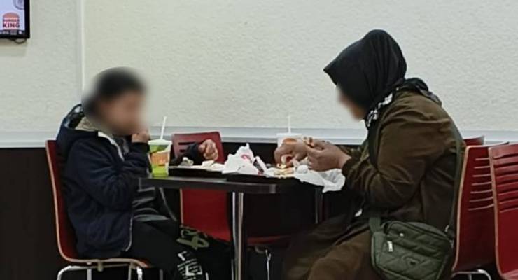 Woman in a hijab and brown coat dining with a child at a fast-food restaurant, drinks and napkins on the table.