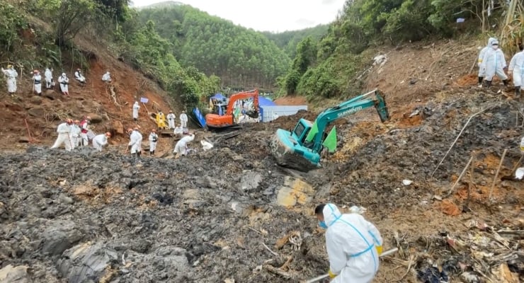 Group of people in white protective suits and masks work at a muddy landslide site with two excavators on a forested hillside.