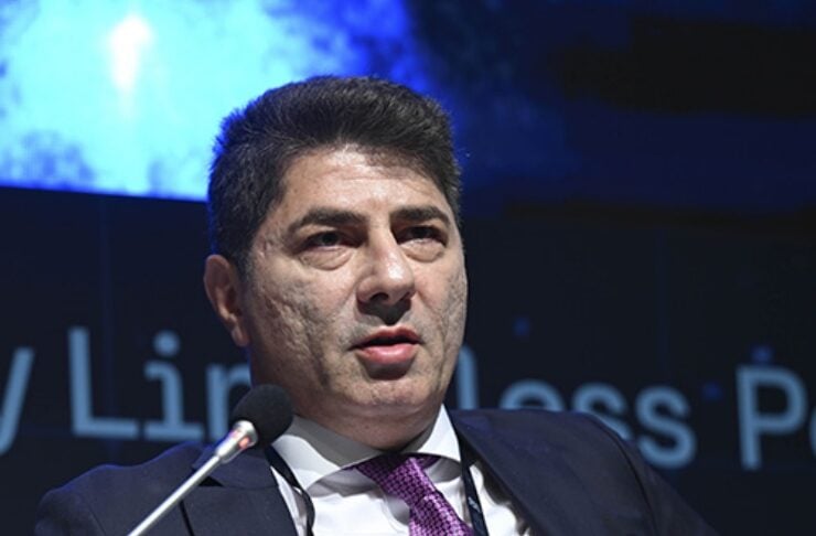 Man in a navy suit with a pink tie speaks at a panel, microphone in front, conference badge hanging from neck