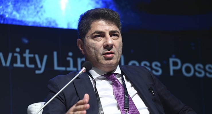 Man in a dark suit and purple tie speaks into a microphone on stage during a panel discussion, with a blurred backdrop.
