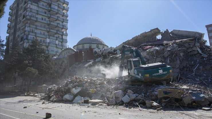 Rubble-filled demolition site with a green excavator perched on debris and a dome-roofed building in the background; tall apartment blocks nearby.