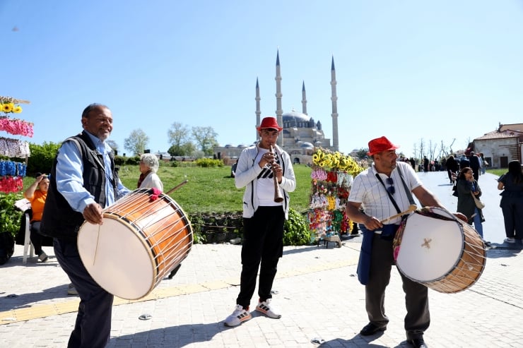 Three street musicians perform in a sunny plaza: two drummers flanking a clarinet player, with a mosque in the background.