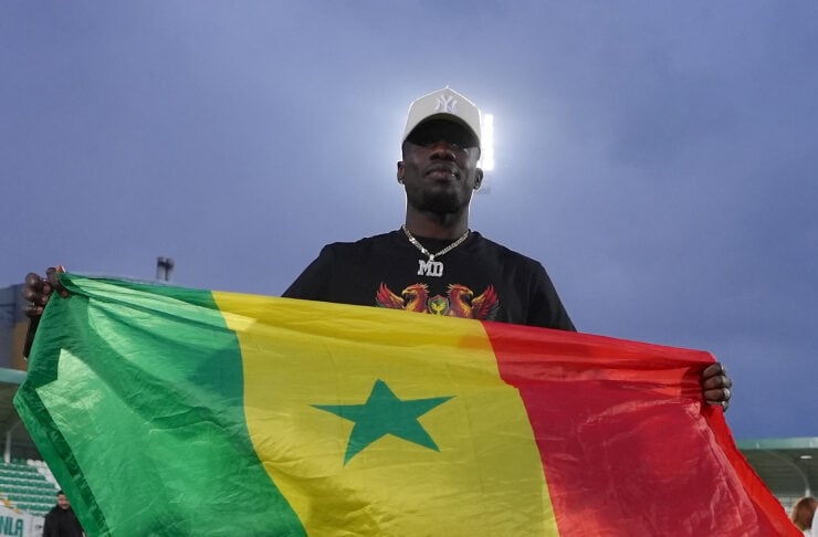 Man on a stadium field holding a large Senegal flag (green, yellow, red with green star). Card-cap and dark shirt visible in the background.