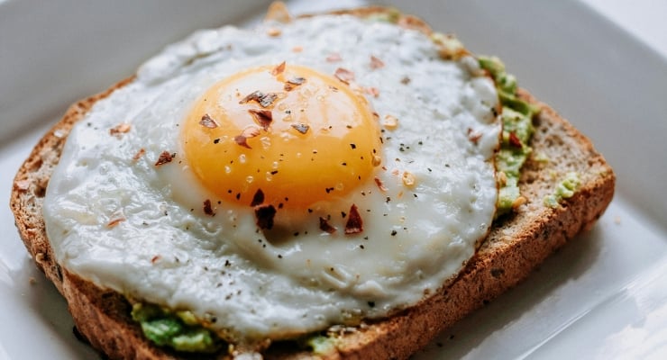 Sunny-side-up egg on avocado toast, sprinkled with red pepper flakes on whole-grain bread.