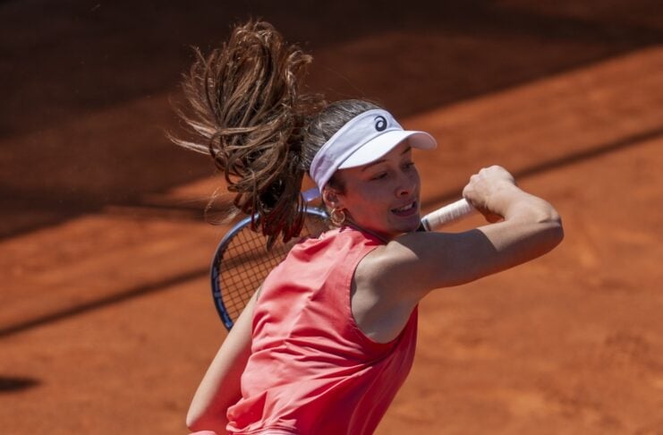 Female tennis player in coral outfit swings a backhand on a red clay court, ponytail flying behind her.