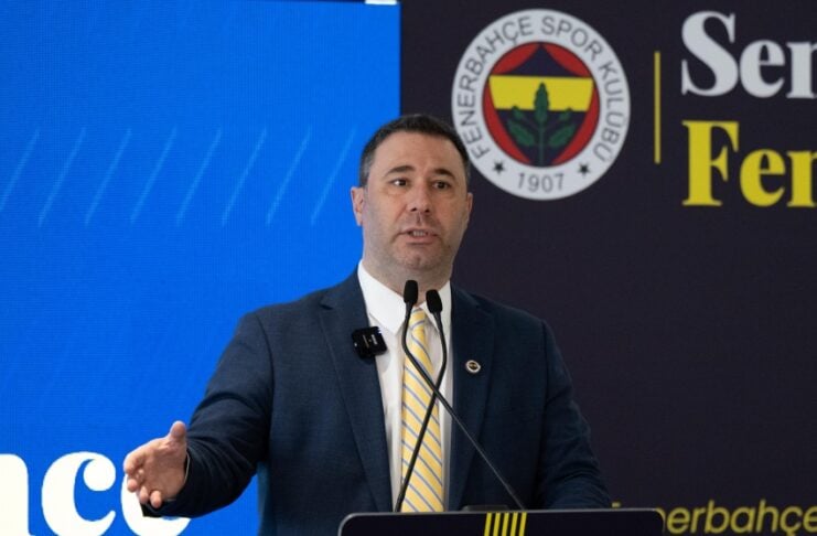 Man in a suit delivering a speech at a podium with microphones; Fenerbahçe logo and event banner behind him.