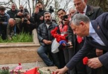 People gather at a stone memorial to lay red carnations; a man in a suit places a flower as others photograph the scene.
