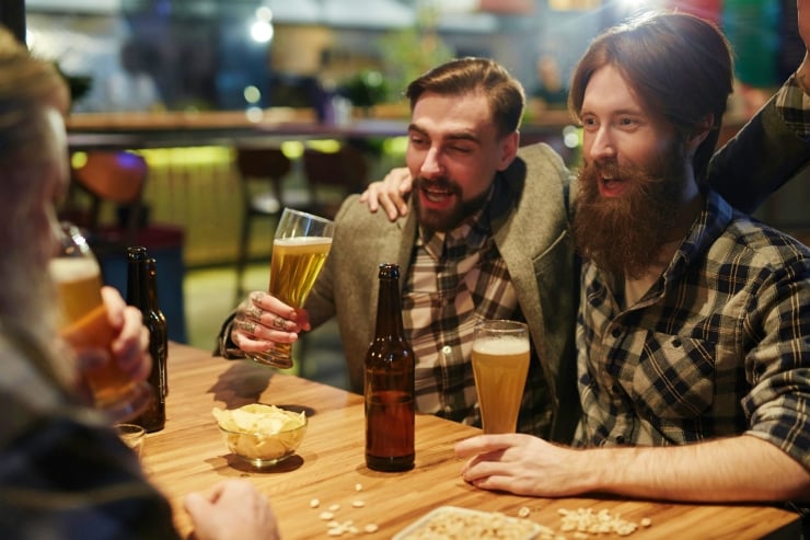 Three friends raising beer glasses and laughing at a wooden table in a lively bar, with snacks nearby.