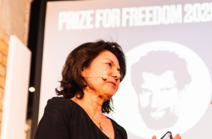 Woman on stage in a black dress, holding a glass award with a headset mic, against a banner reading 'PRIZE FOR FREEDOM'.