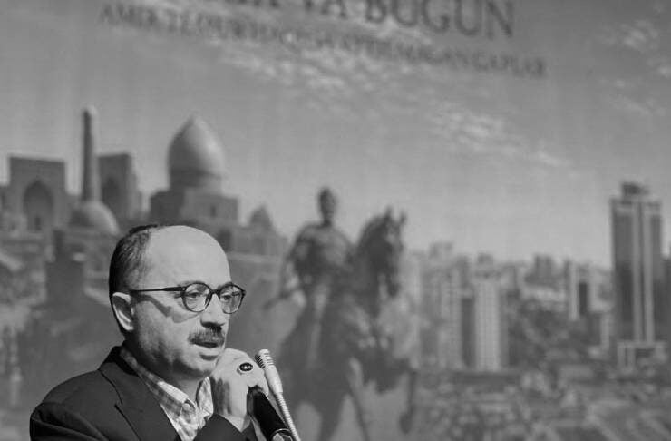 Man wearing glasses speaks into a microphone at a clear podium during a formal event; cityscape backdrop and Bursa signage visible behind him.