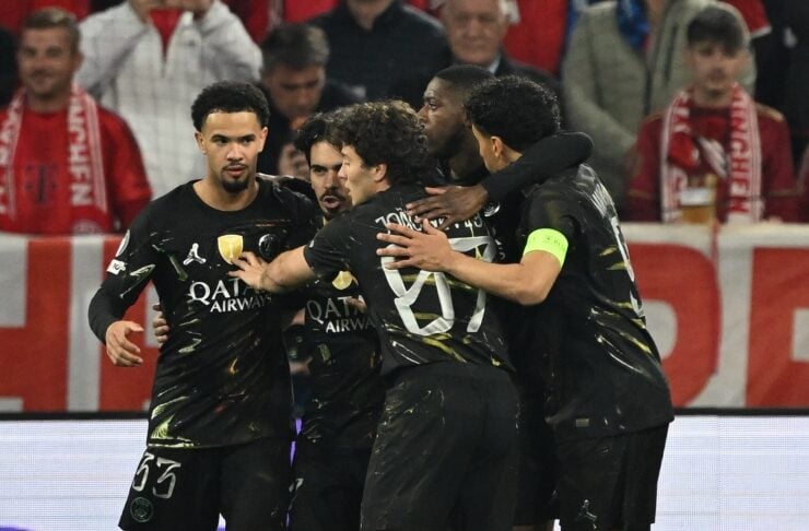 PSG soccer players in black kits celebrate a goal, arms around each other on the pitch, one with a yellow captain's armband.