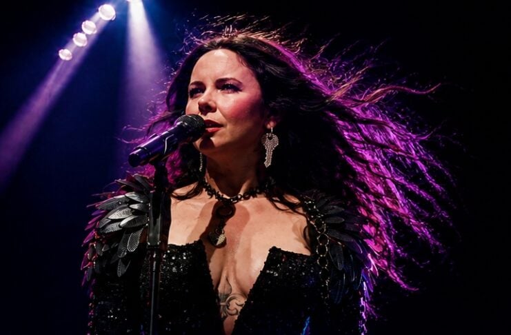 Female singer performing on stage under purple spotlights, wearing a black sequined outfit and statement jewelry, singing into a microphone while a cheering crowd watches.