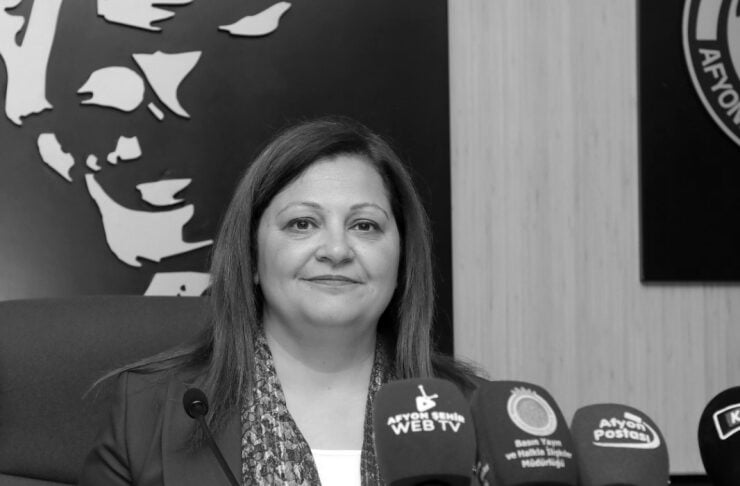 Woman seated at a press conference speaking to reporters, with multiple microphones in front of her.