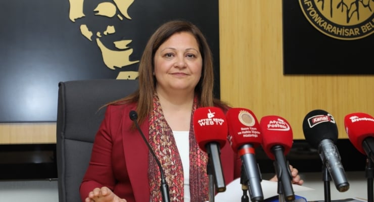 Woman in a red blazer at a press conference, seated behind microphones.