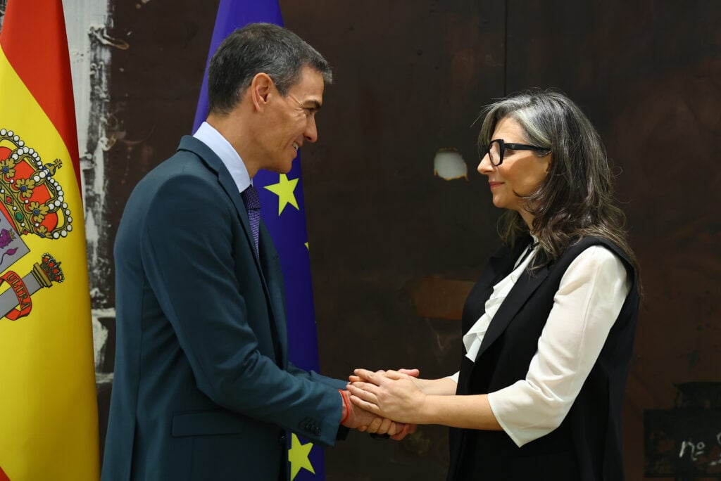 Man in a suit and a woman with glasses shake hands, with the Spanish and European flags in the background.