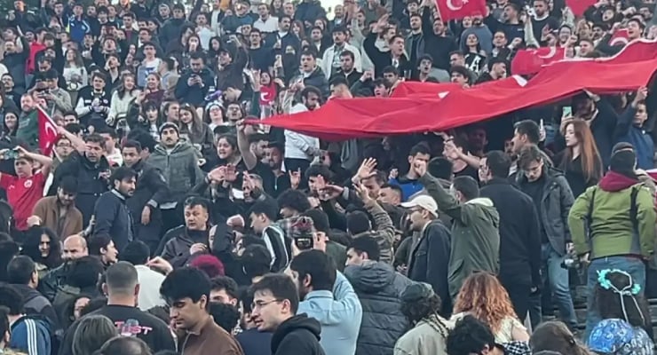 Large crowd at a rally with many Turkish flags and a long red banner sweeping overhead versus the crowd.