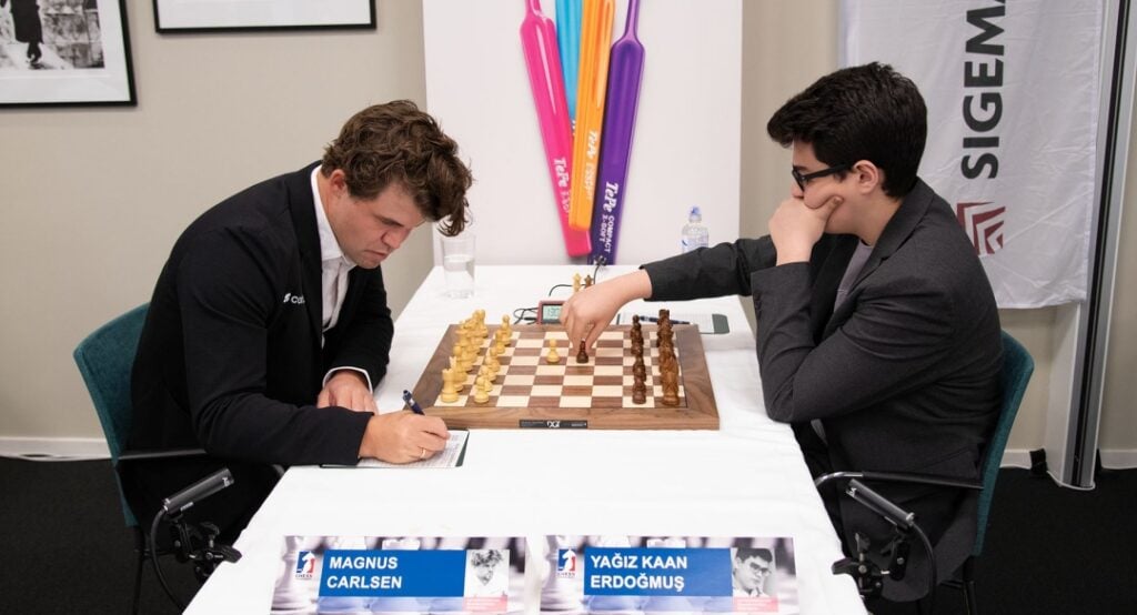 Two men in suits play chess at a tournament table; one signs a score sheet while the other watches thoughtfully.